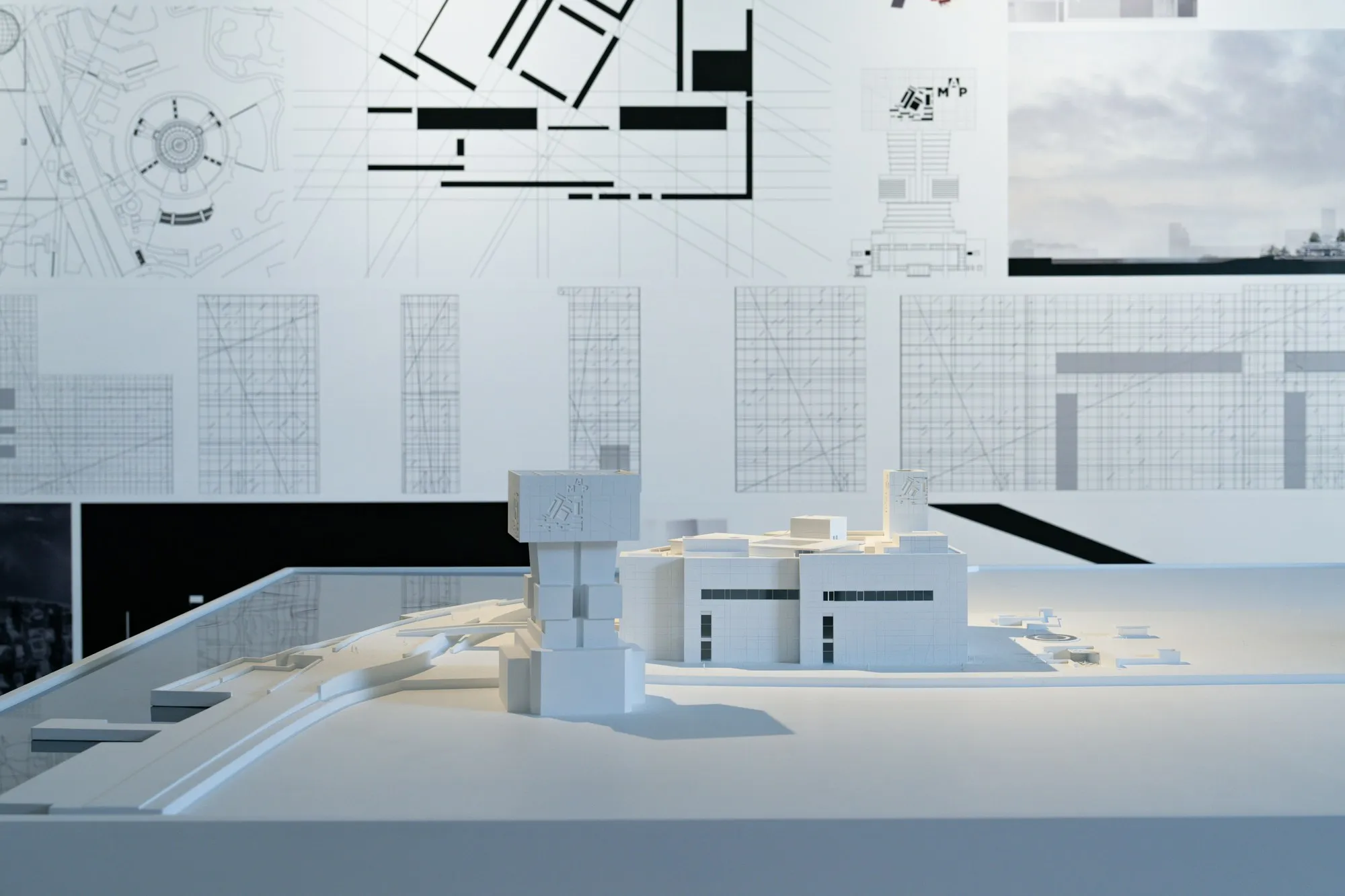 White architectural site model beneath plans and elevations hung on a gallery wall