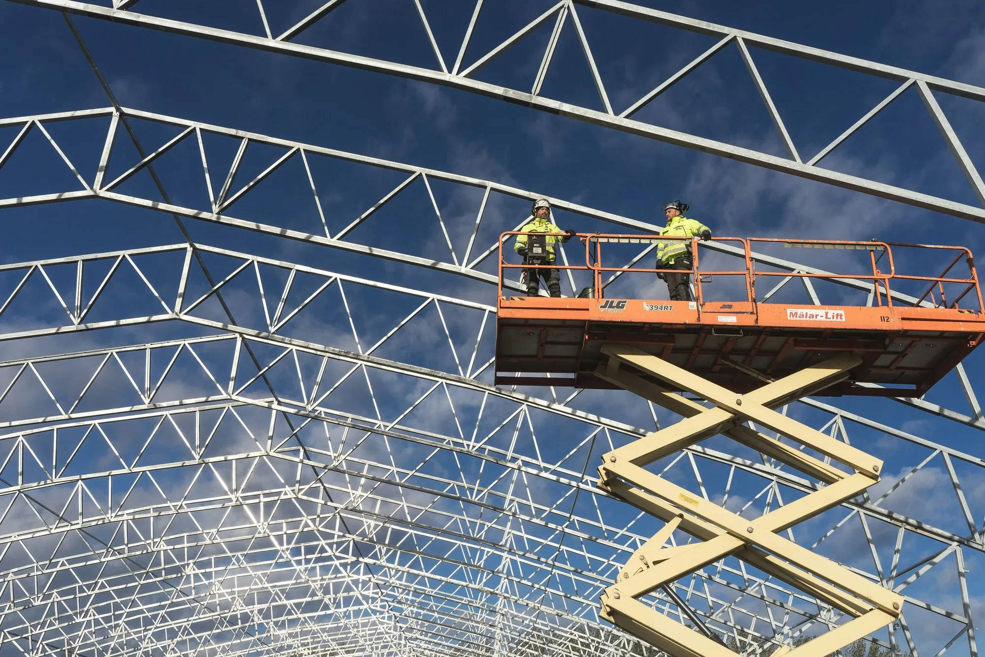 Two site operatives on a lift under a steel roof structure