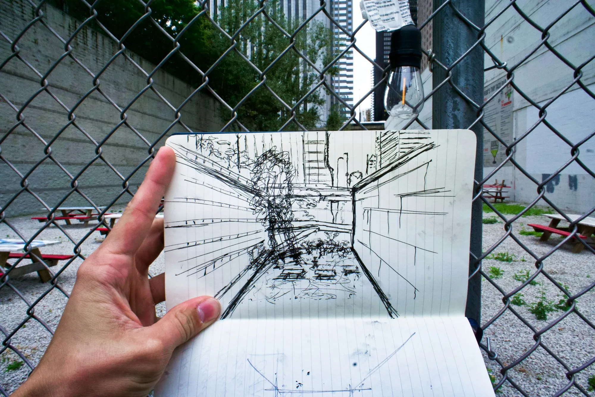 Hand holding a perspective sketch of a building beyond a fence