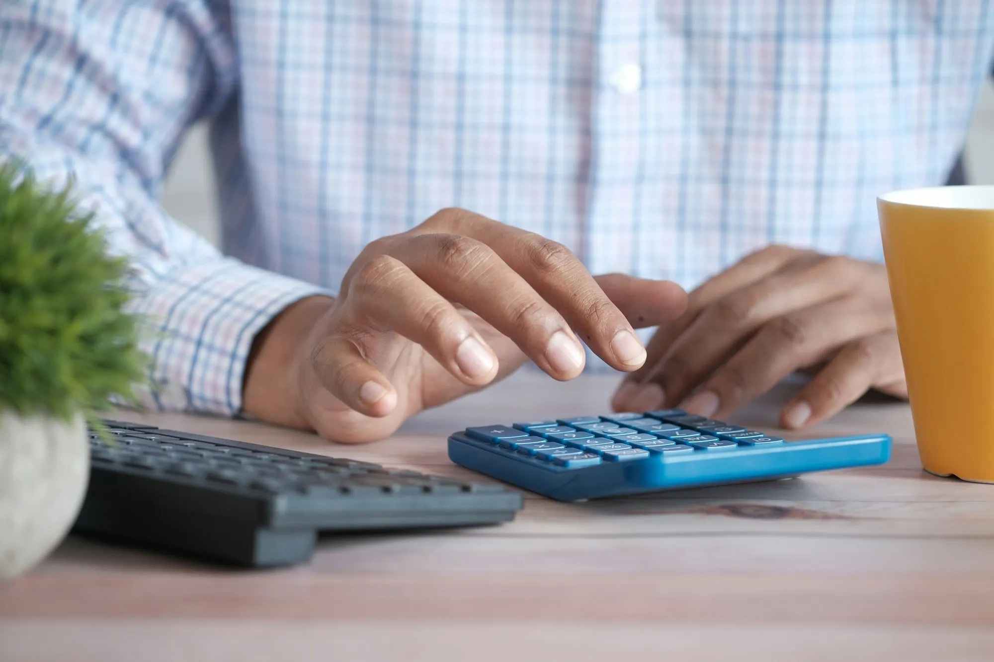 Hands using a calculator at a desk beside a keyboard (cost-planning feel)