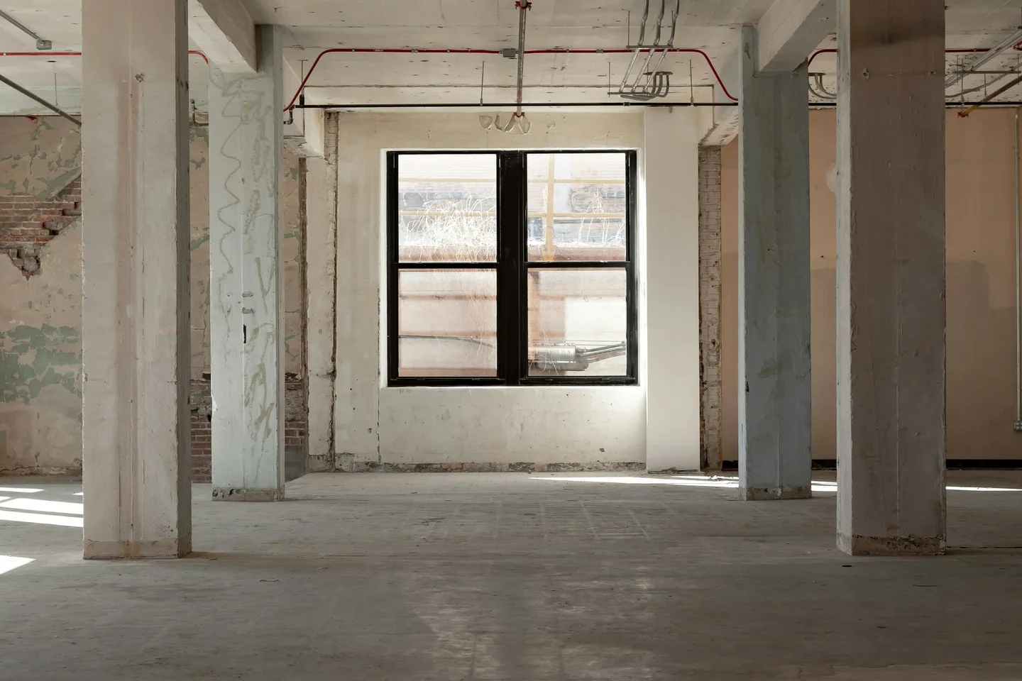 Stripped-out industrial building interior with concrete columns and a tall black-framed window in daylight