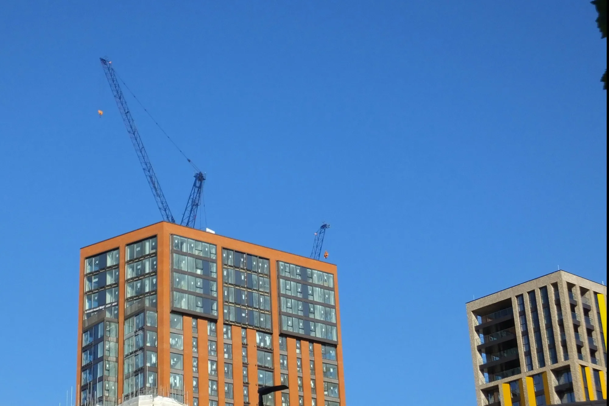Brick-clad residential tower near completion with tower cranes against a clear blue sky, East London