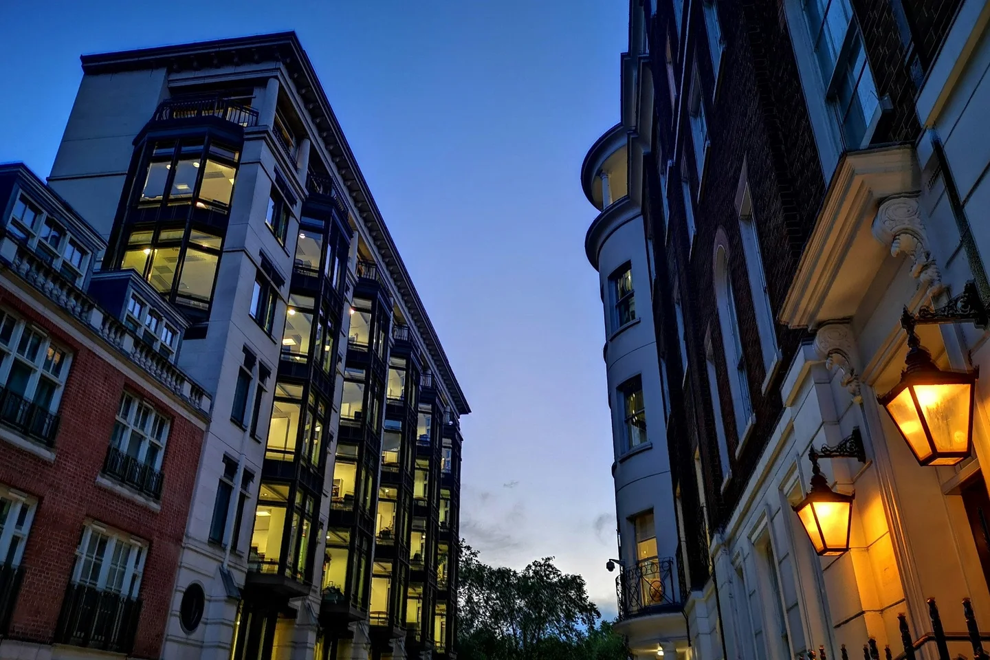 London street at dusk with lit office and residential windows between brick and stone buildings, gas-style lamps glowing