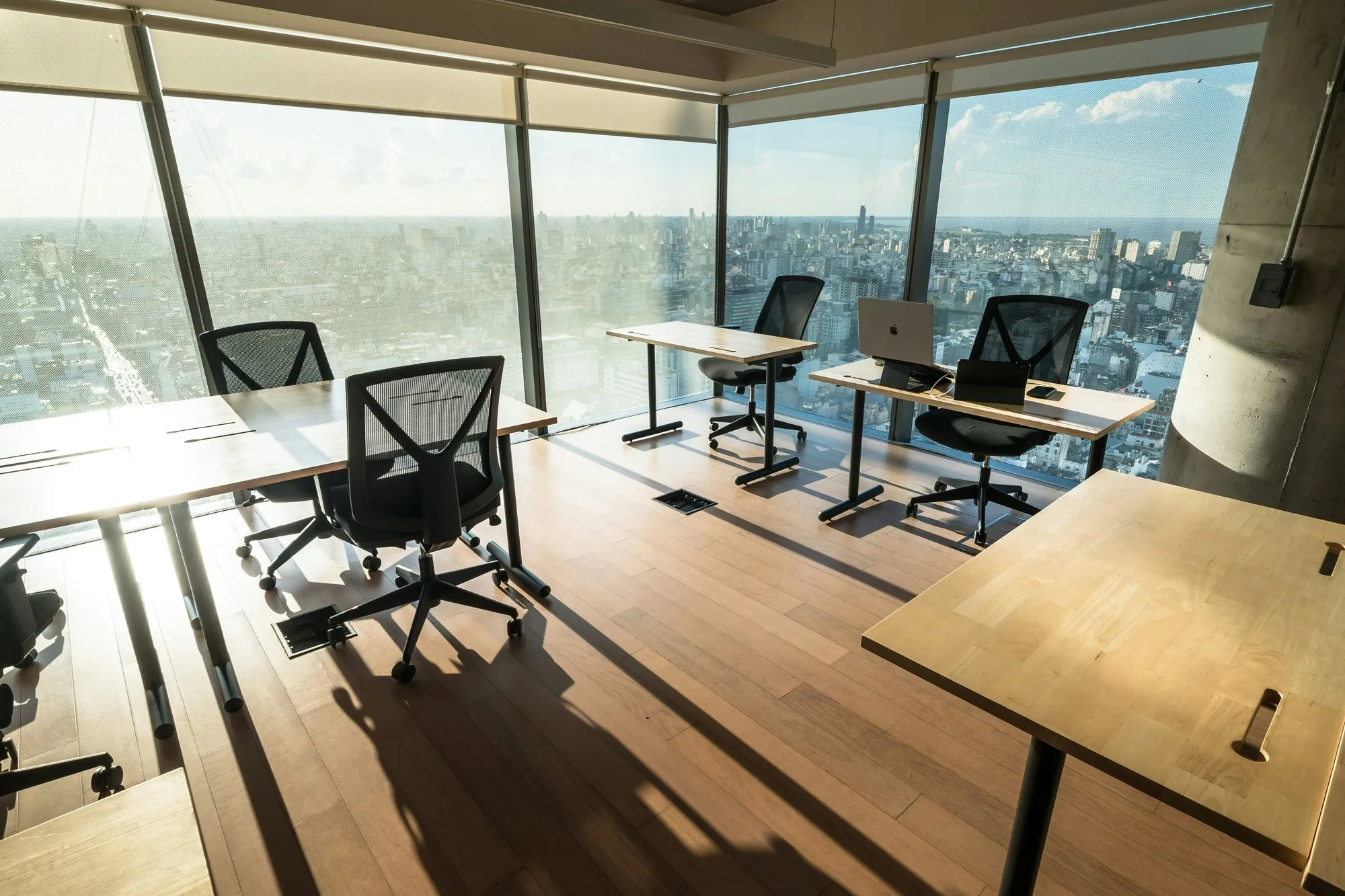 Sunlit office floor with timber desks and mesh chairs beside floor-to-ceiling glazing over a city