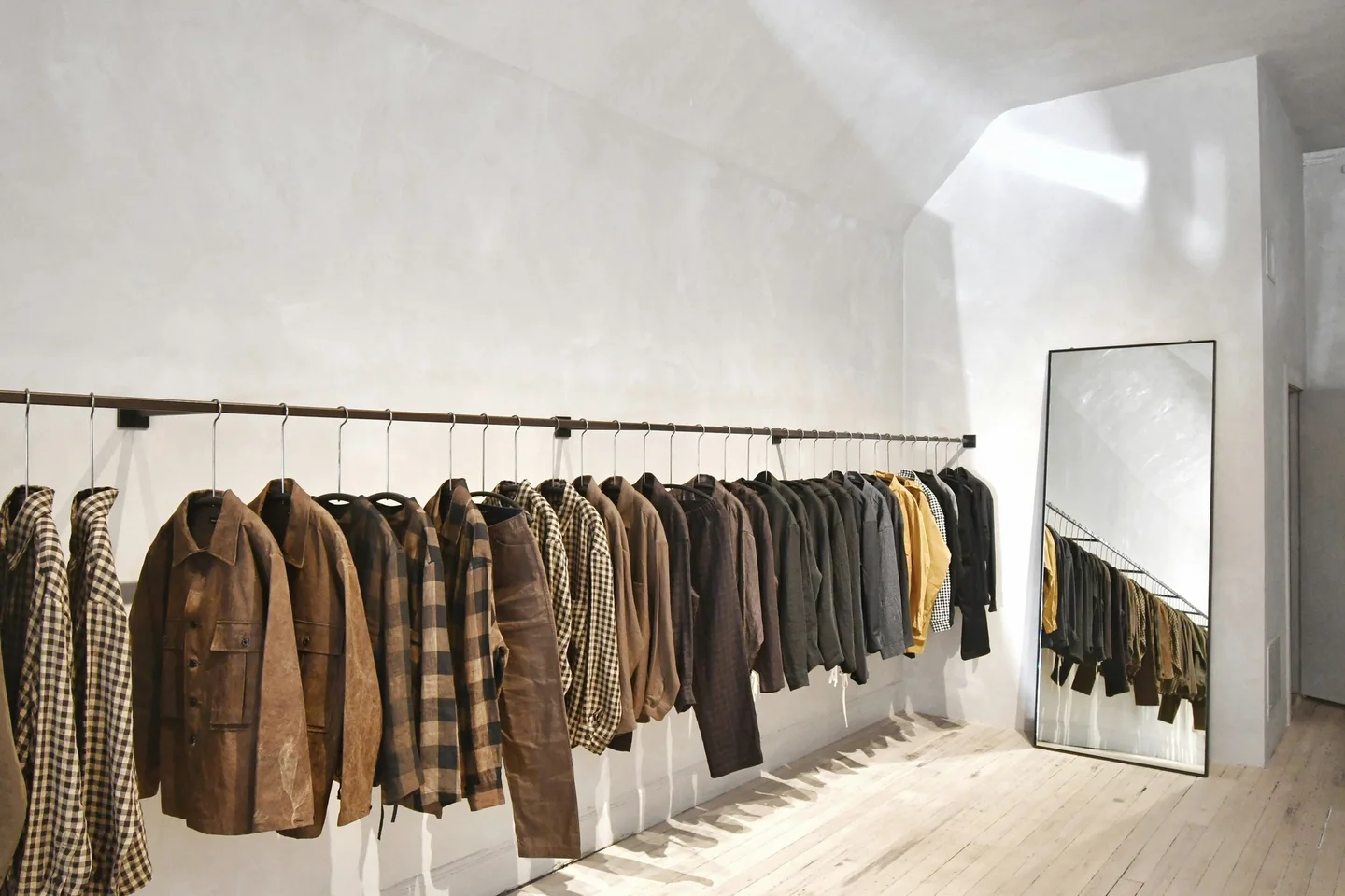 Boutique rail of jackets and knitwear on a plastered wall beside a floor mirror, pale timber floor