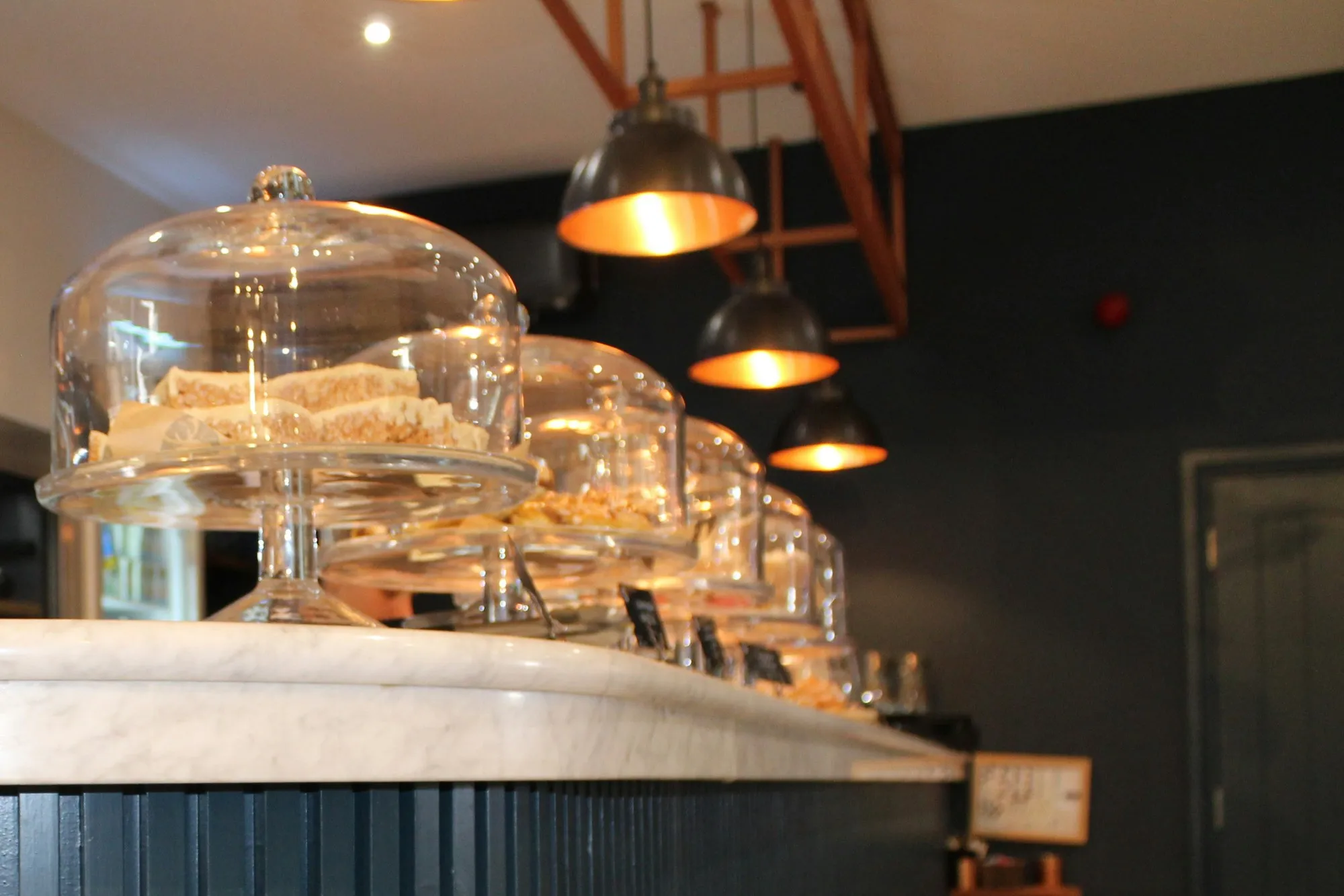 Marble-topped cafe counter with glass cake domes and industrial pendant lights, fluted painted front