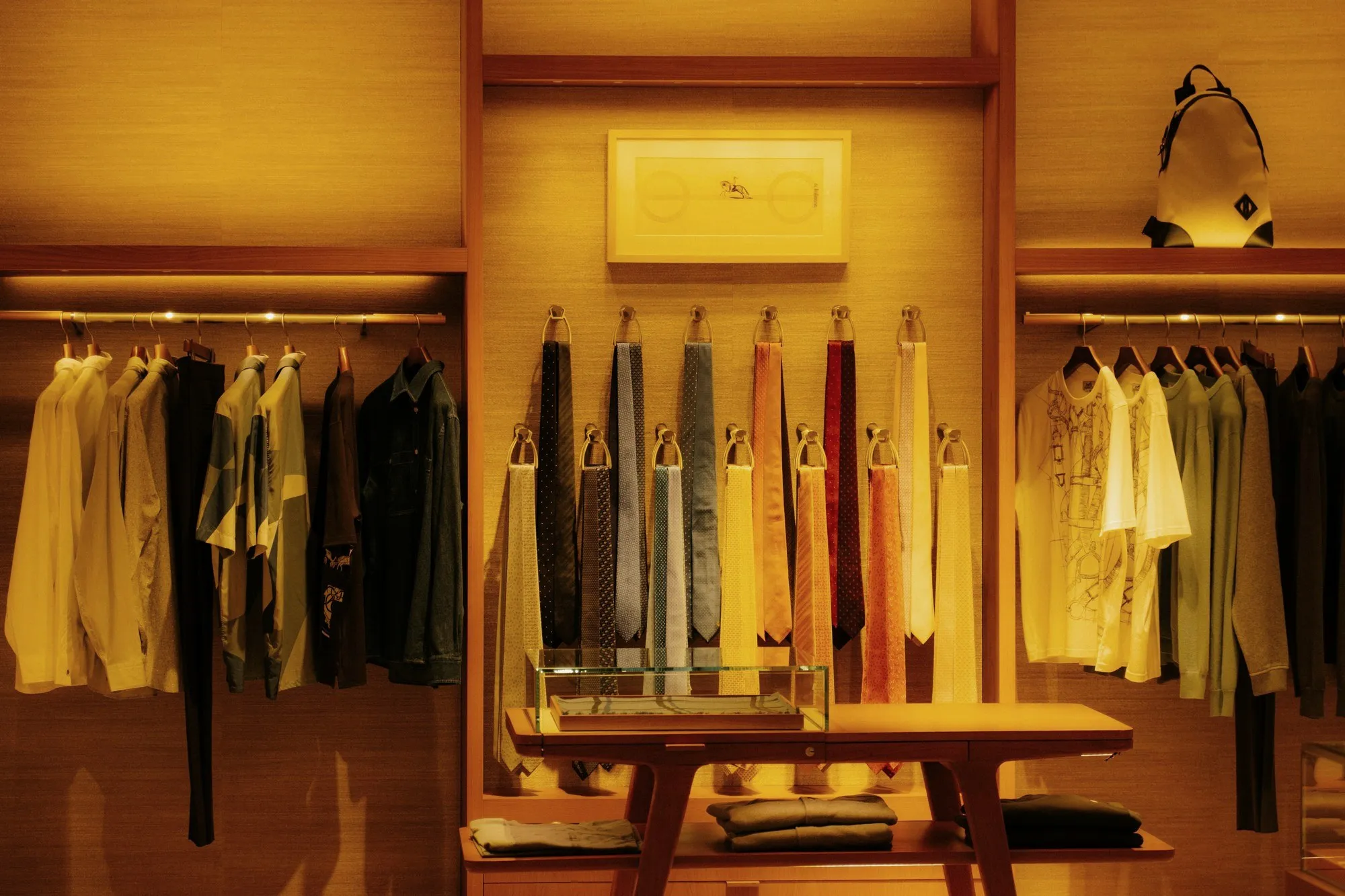 Warm-lit fashion boutique wall with rails, shirts, ties and a timber display table