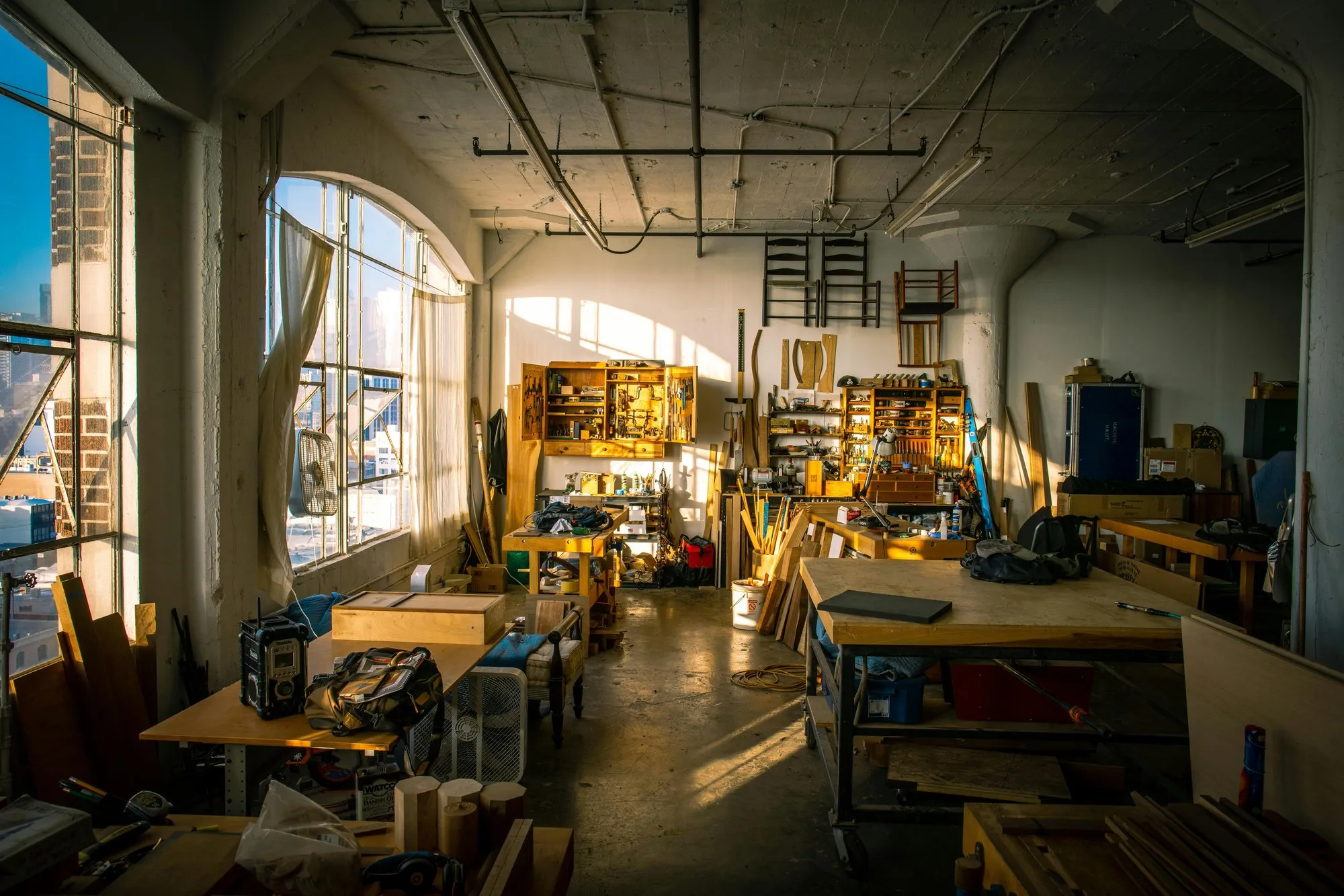Sunlit joinery workshop with benches, timber stock and tool wall in an industrial loft