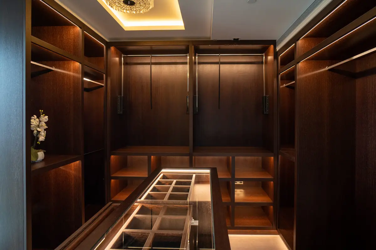 Dressing room with dark wood wardrobes, warm strip lighting and a glass-topped island in a Kensington Olympia penthouse