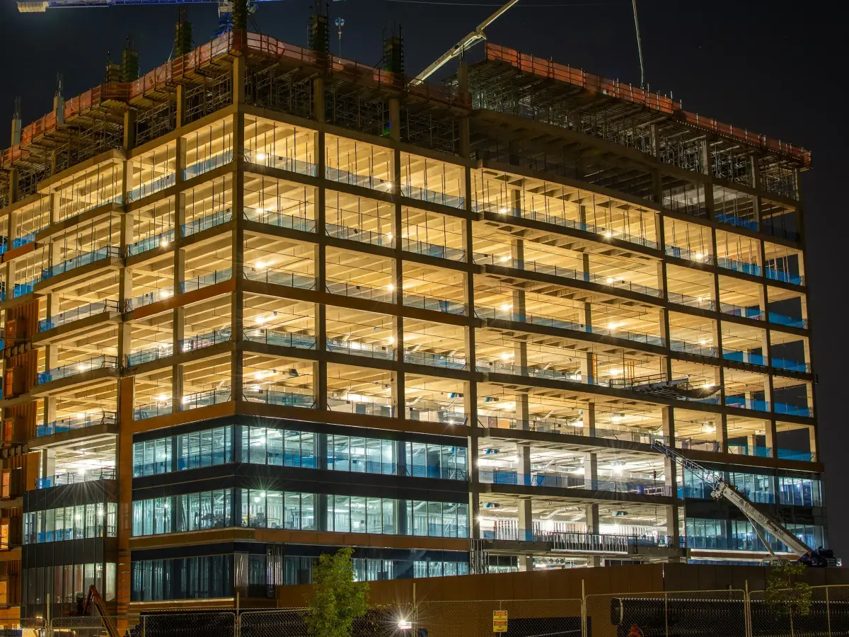 Multi-storey office building under construction at night, its lit concrete frame topped by a tower crane
