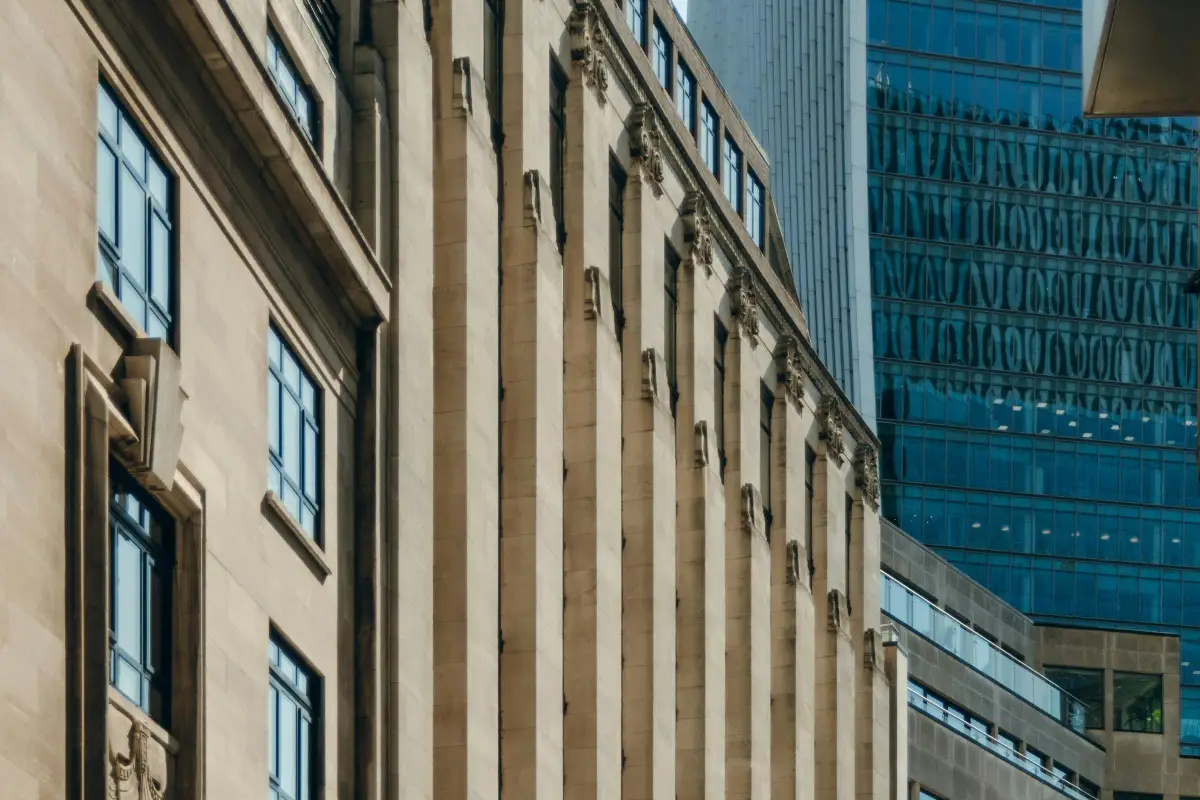 Stone-fronted office building in the City of London beside a glass tower reflecting the sky