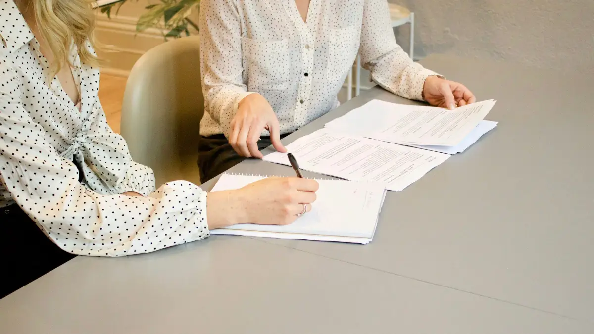 Two people at a table reviewing and signing contract documents with a pen and notepad