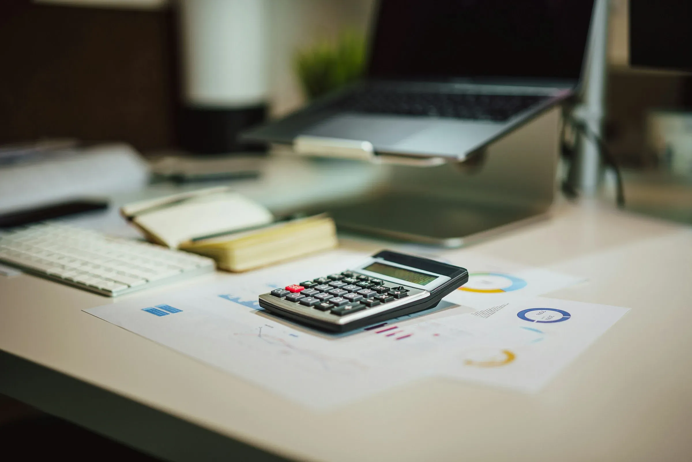 Calculator resting on printed charts on a desk beside a keyboard and laptop