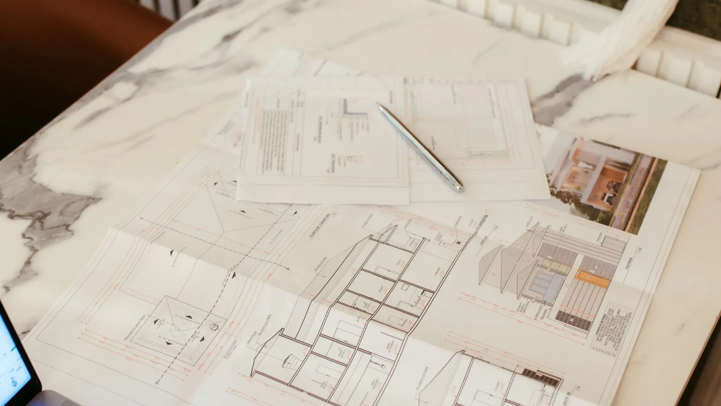 Architectural drawings and a pen on a marble desk beside a laptop