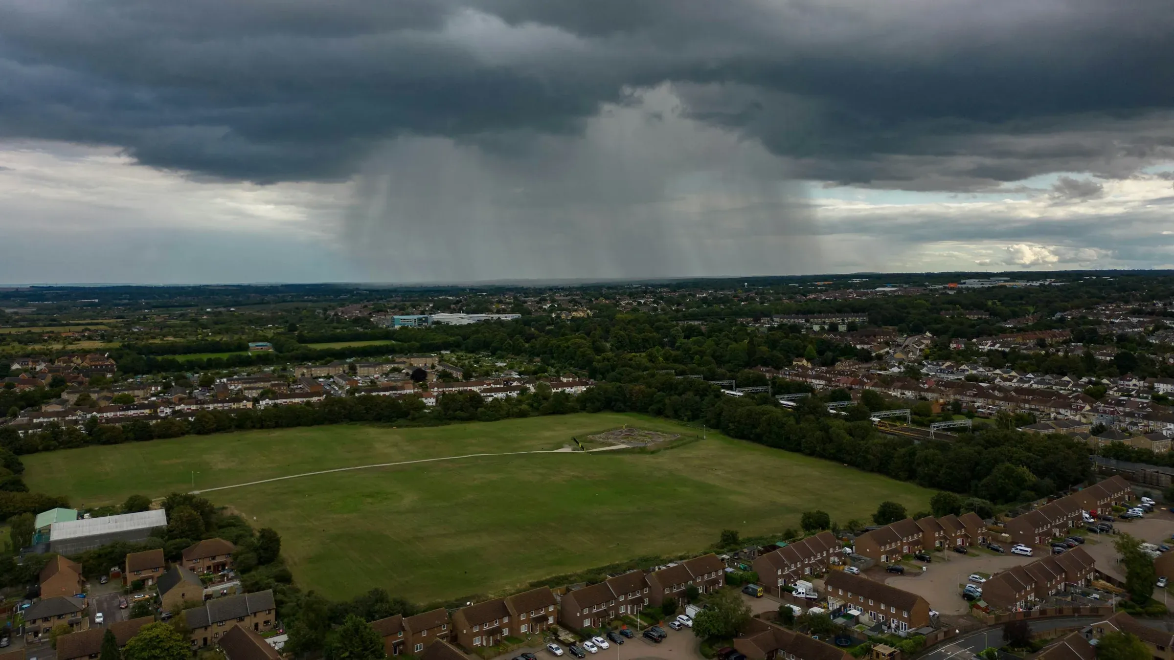 Aerial view of a green field bordered by rows of houses in Medway, Kent, under a rain cloud