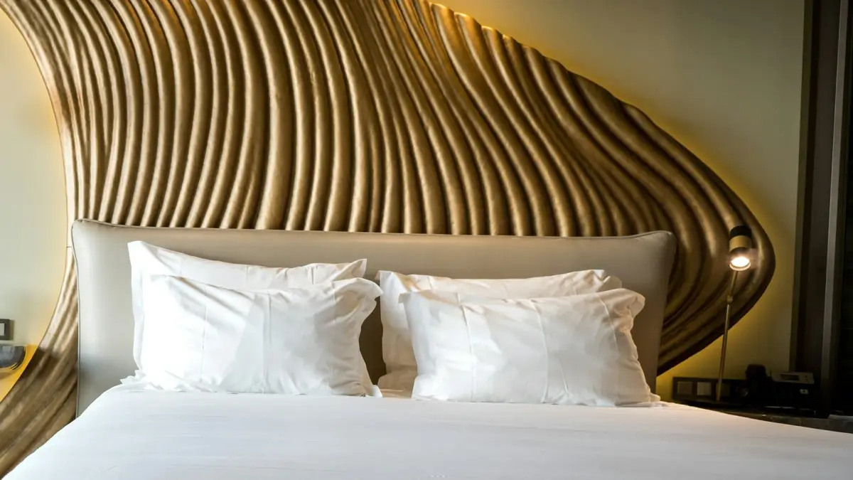 Hotel bedroom with a sculpted gold wall panel behind an upholstered headboard, white bed linen and a bedside reading light