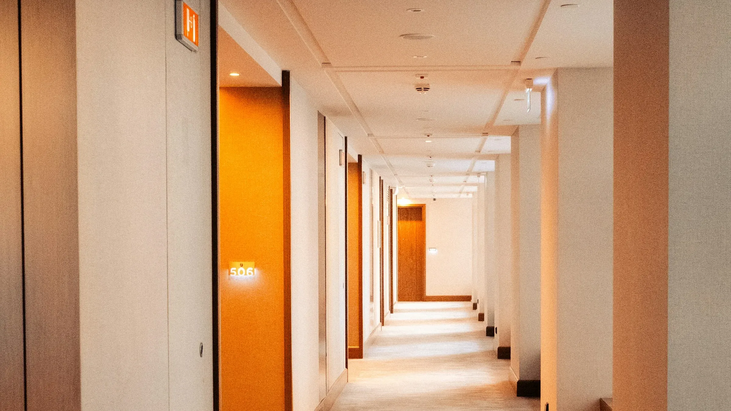 Long hotel guest corridor with warm lighting, carpeted floor and timber-framed bedroom doors