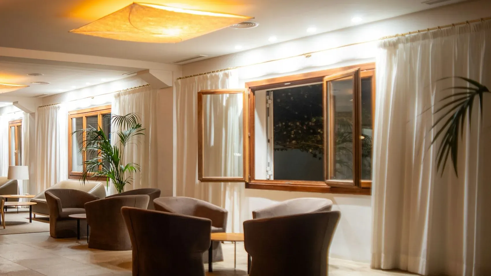 Hotel lounge with upholstered tub chairs around low tables, pendant ceiling lights and white curtains at timber windows