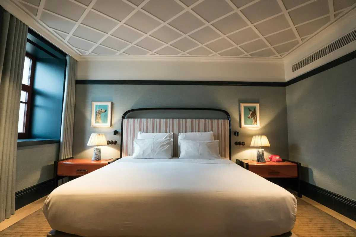Hotel bedroom with a striped upholstered headboard, two lacquered bedside tables with table lamps, framed artworks and full-height curtains