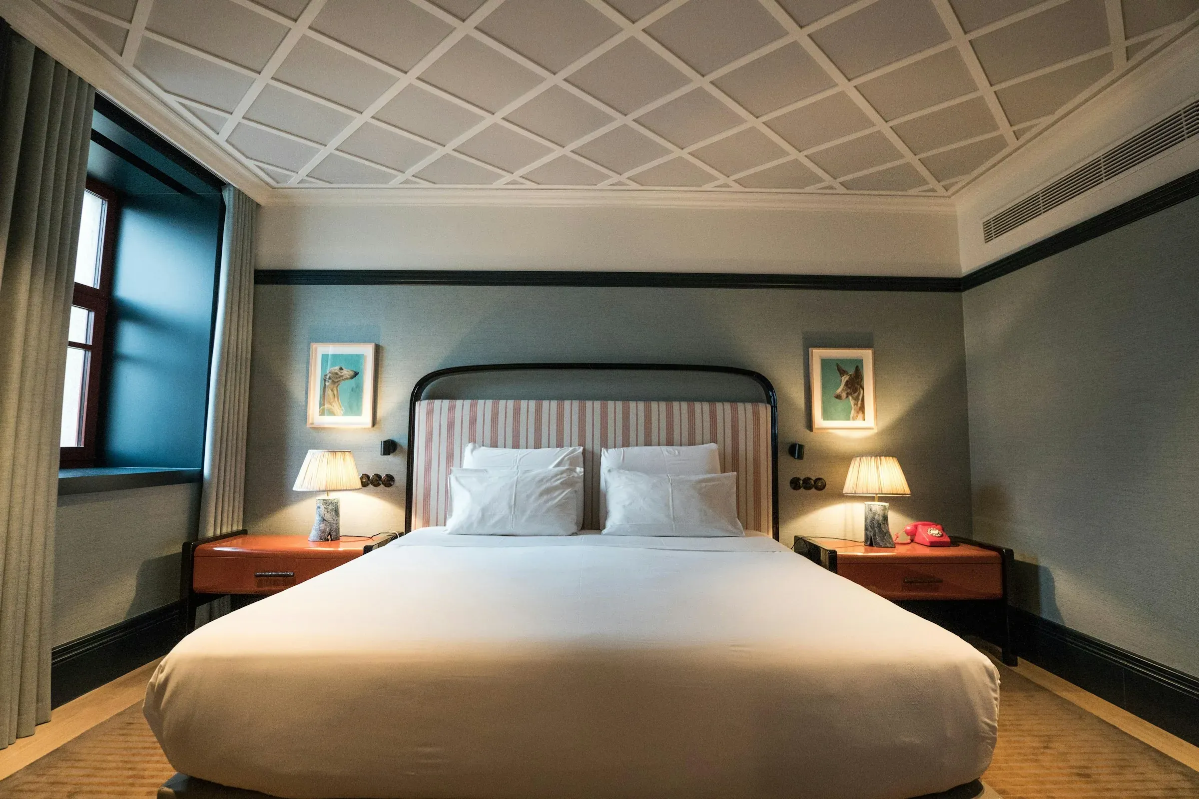 Hotel bedroom with a striped upholstered headboard, two lacquered bedside tables with table lamps, framed artworks and full-height curtains