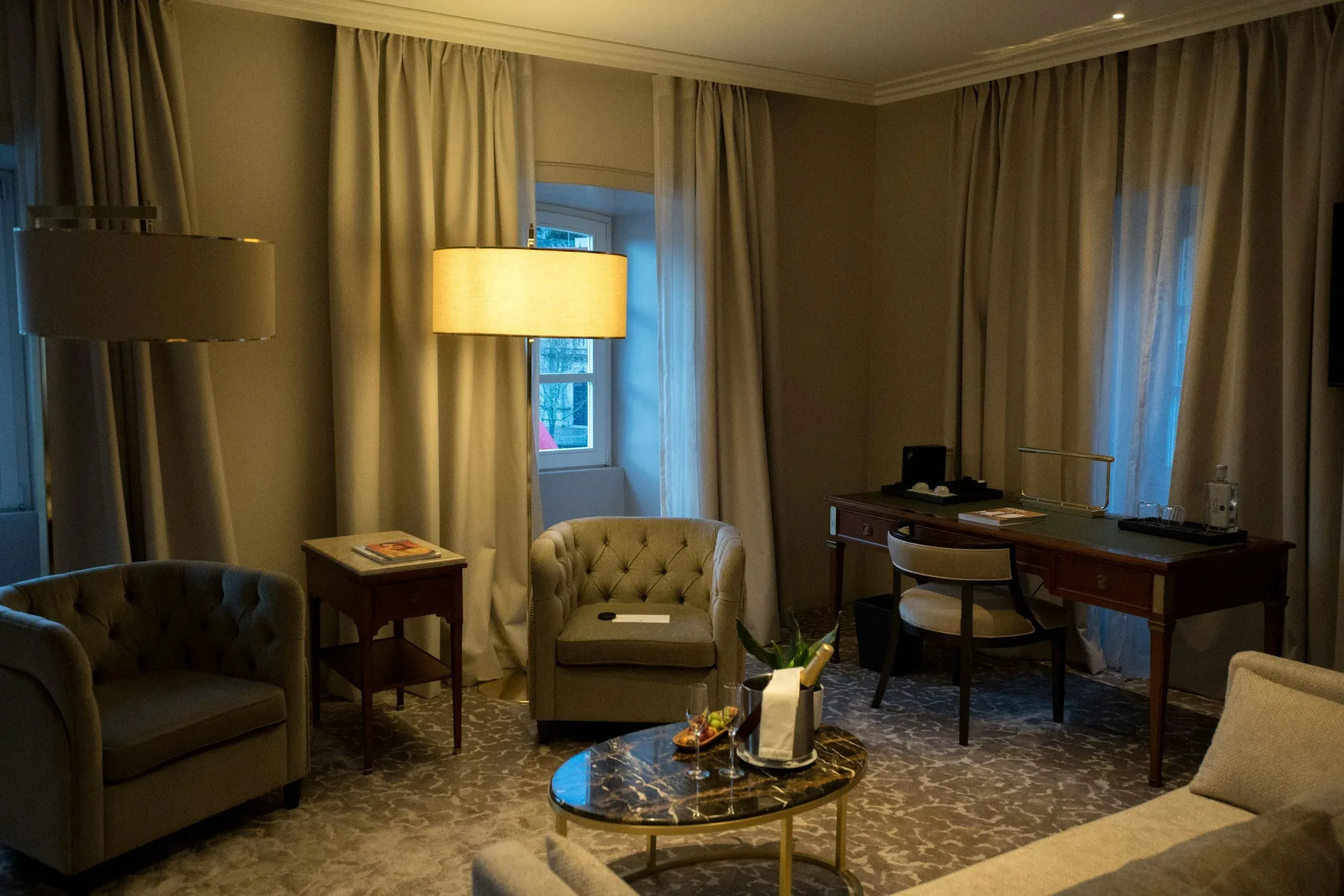 Hotel suite sitting area with tufted armchairs, a marble coffee table, a side table, floor lamps, a writing desk and full-height curtains