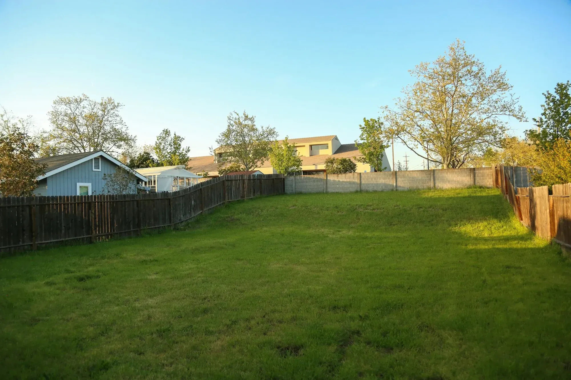 Empty grassed plot of land behind timber fencing, with houses and trees beyond