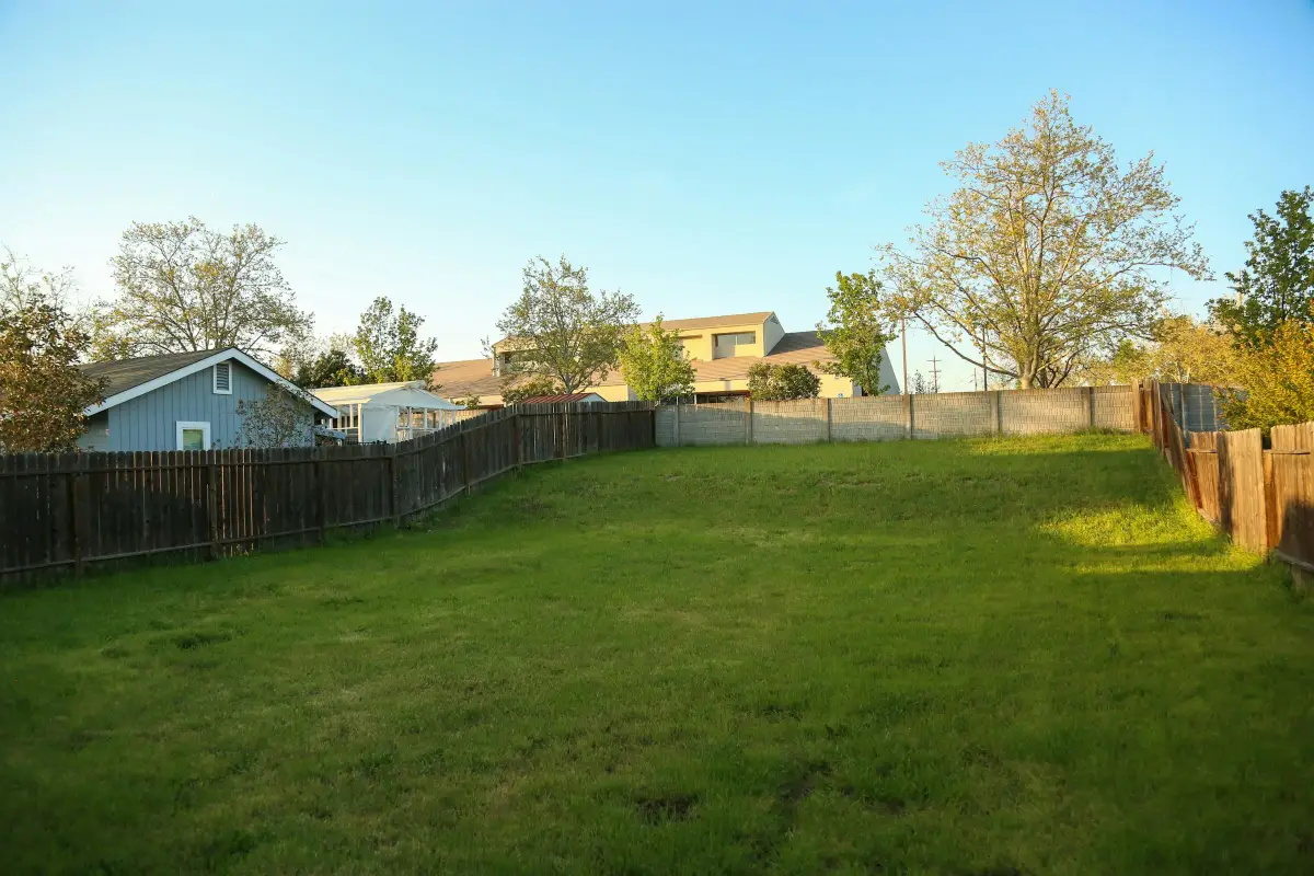 Empty grassed plot of land behind timber fencing, with houses and trees beyond
