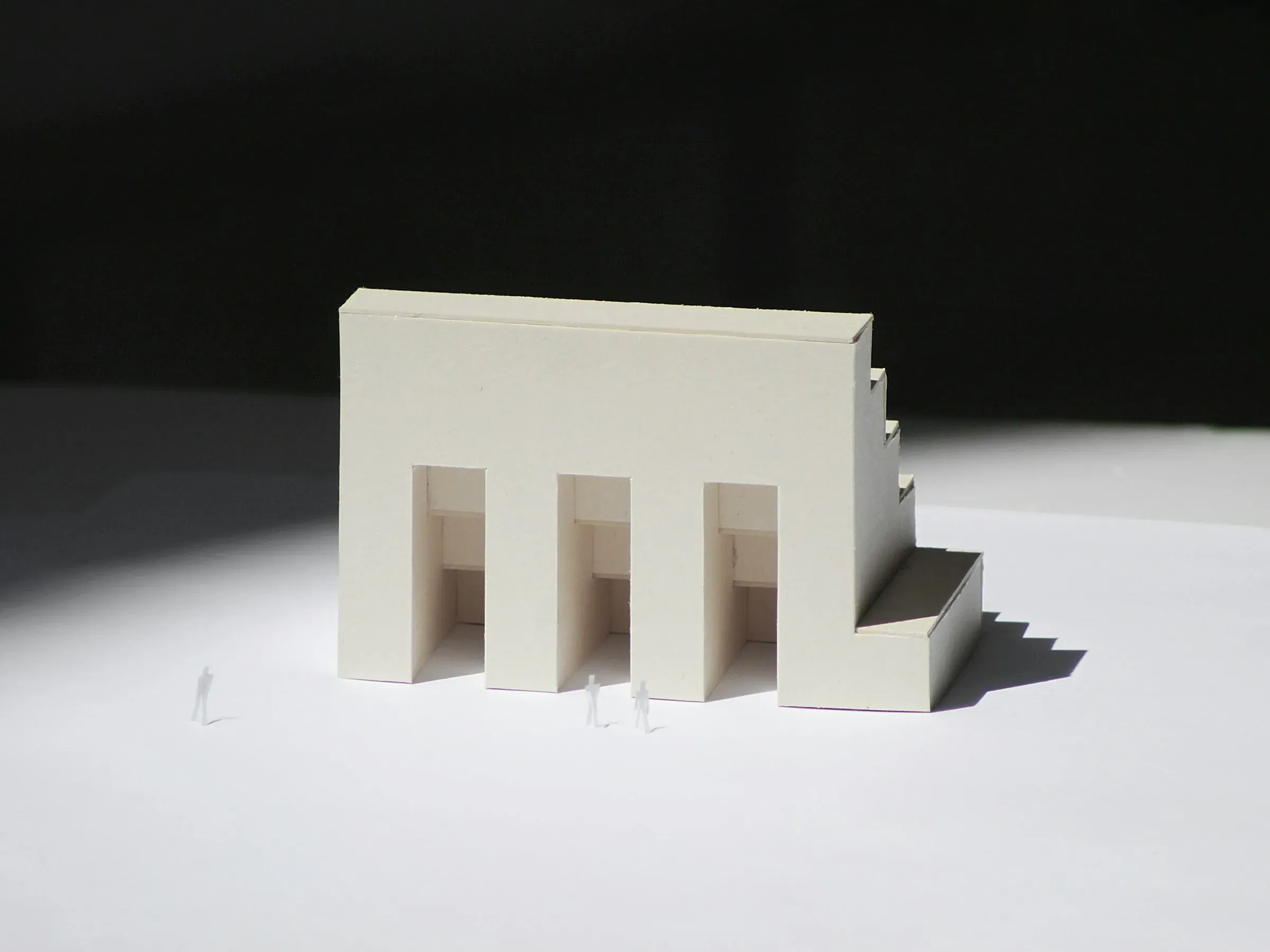 White card concept model of a building with three bays and a stepped roof, lit from the side on a white table