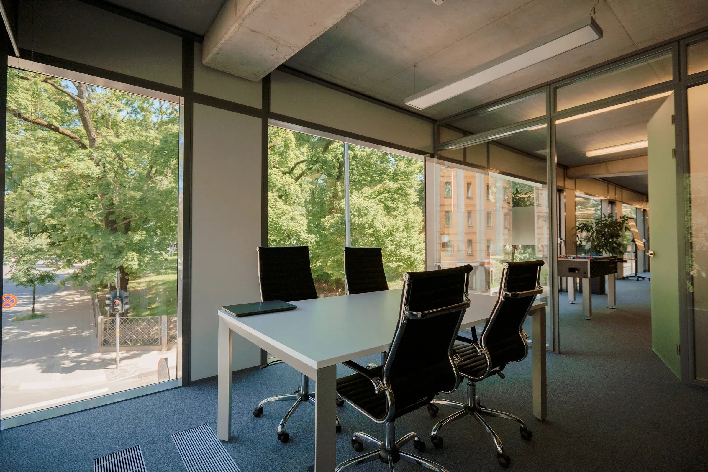 Glass-walled meeting room with a white table and black chairs, with tall windows onto trees