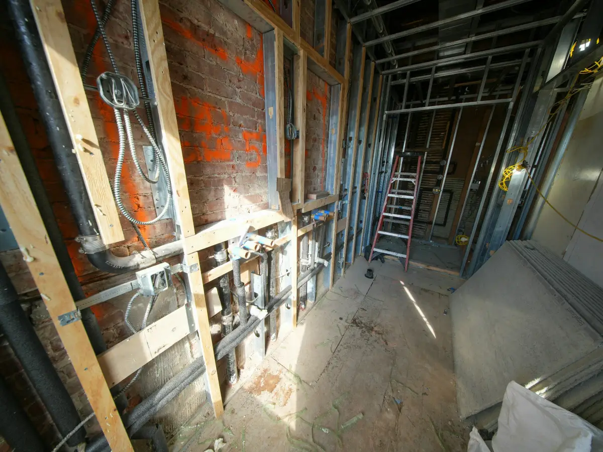 Gutted room mid-renovation with exposed brick, new pipework, timber and metal stud framing and a stepladder