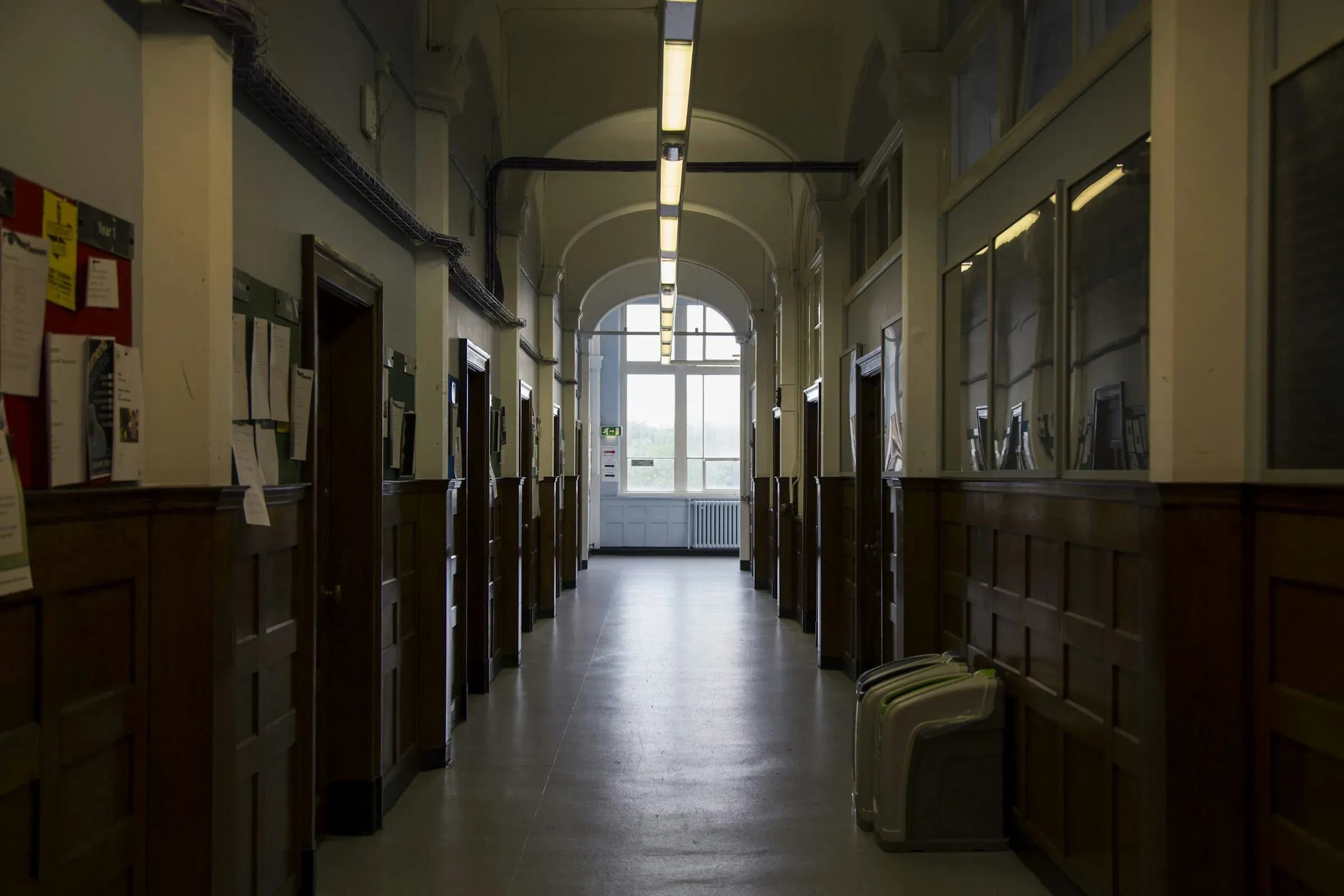 Long Victorian school corridor with panelled walls, noticeboards and a tall arched window at the end