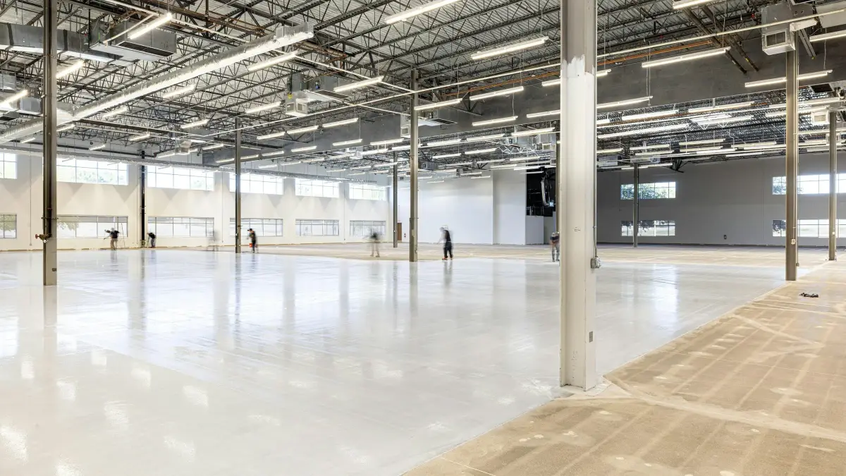 Large empty industrial hall with a white resin floor, steel columns, roof trusses and high windows