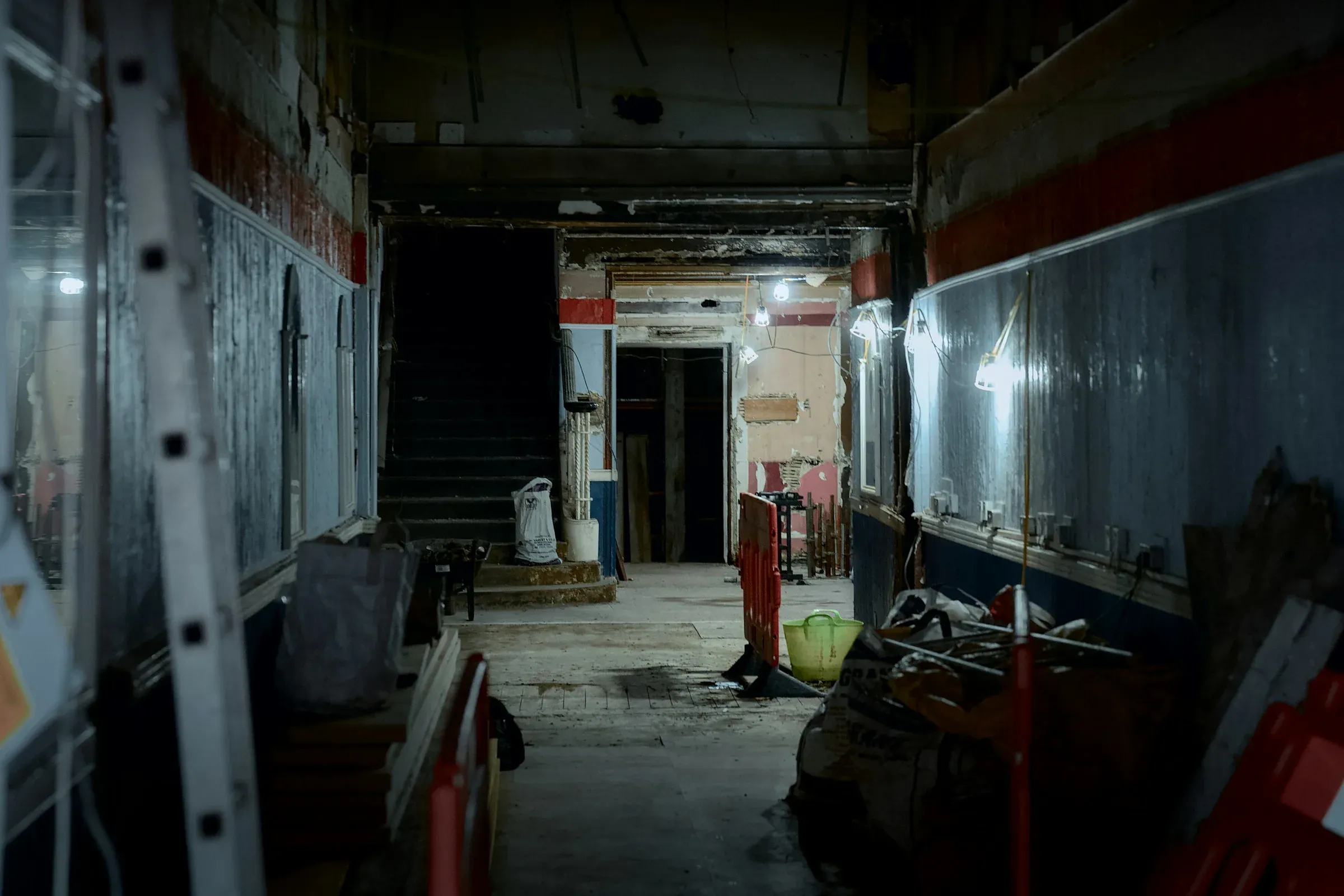 Empty stripped-out corridor on a building site with temporary lights, barriers, a ladder and bagged materials left in place
