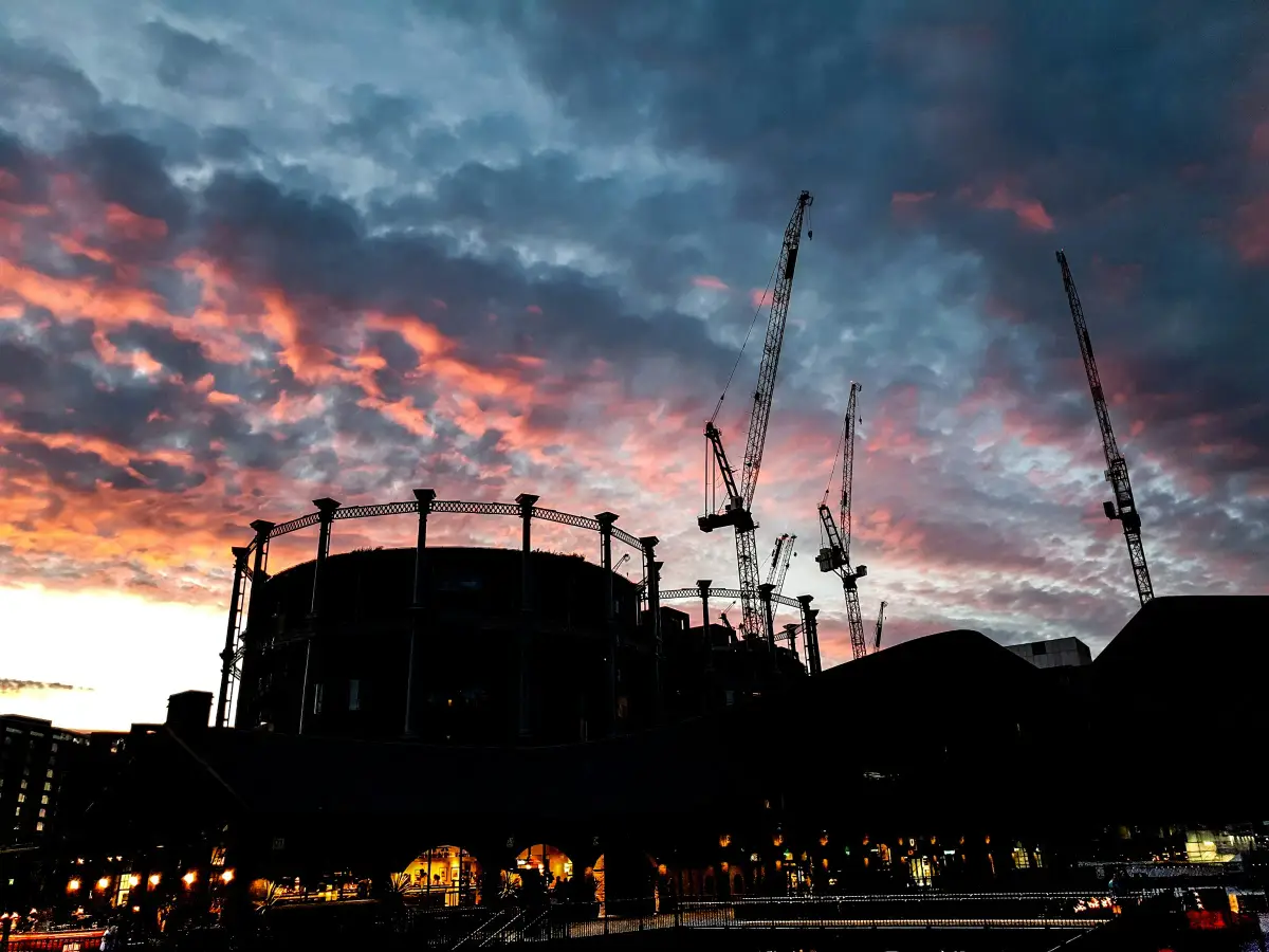 Tower cranes and the frame of a Victorian gasholder silhouetted against a red sunset over a London regeneration site