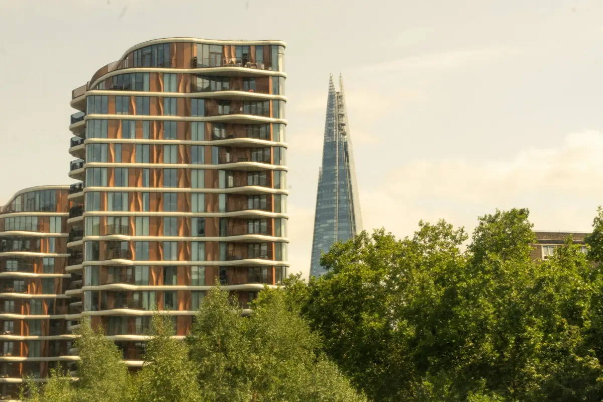 Curved modern residential tower with balconies and bronze-toned cladding beside The Shard, seen through trees in London