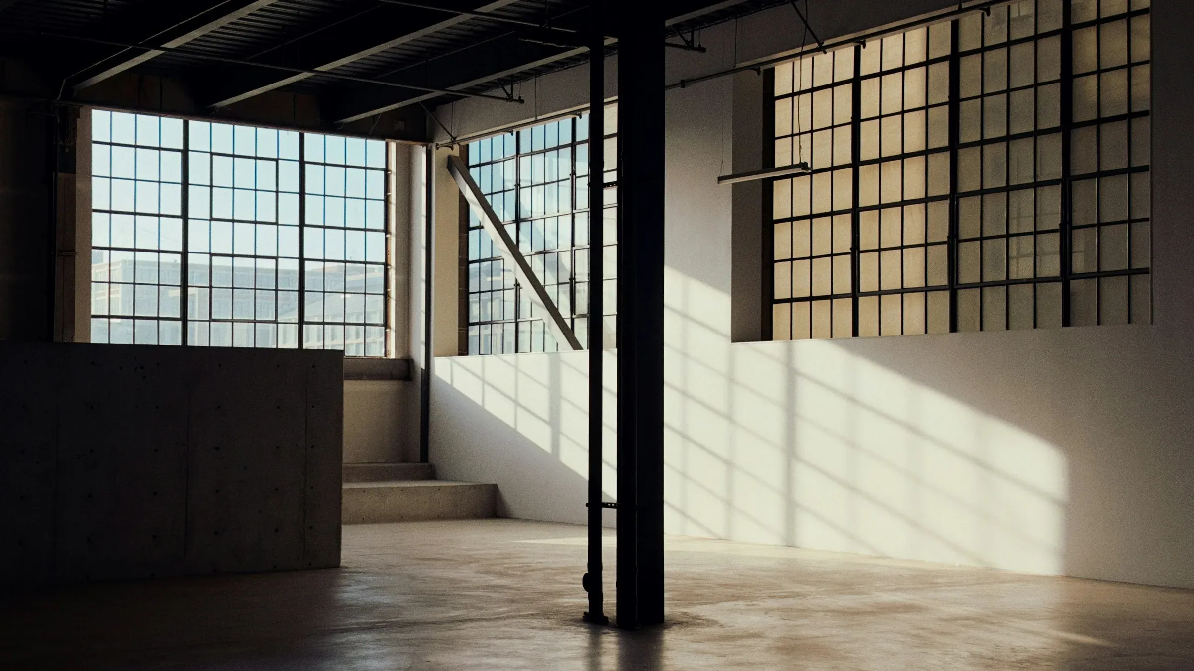 Empty industrial building with tall steel-framed windows, a steel column and a concrete floor lit by low sun